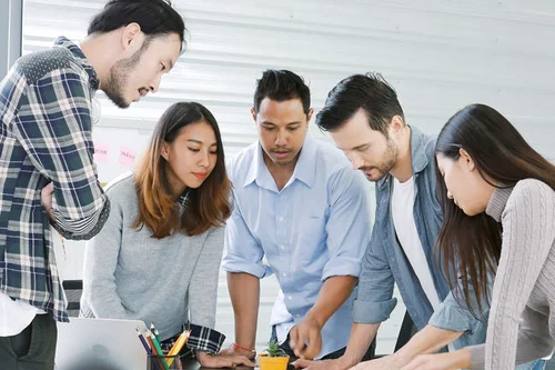 A group of 5 people talking over a desk. They are collaborating as a team. 